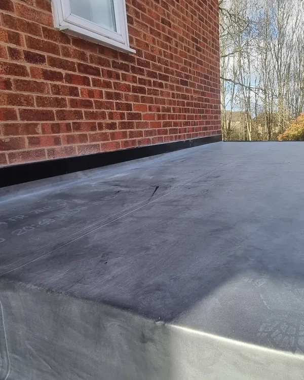 A black EPDM rubber membrane covering a flat roof on a brick extension, sealed along the wall with a trim, trees and a garden beyond