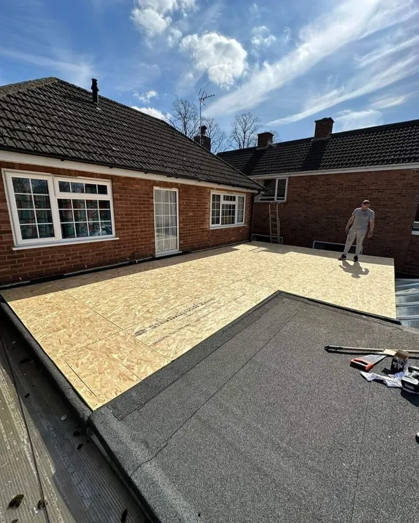 A large flat roof beside a brick bungalow, part covered in new timber deck boards and part in mineral felt, with a roofer standing on the deck
