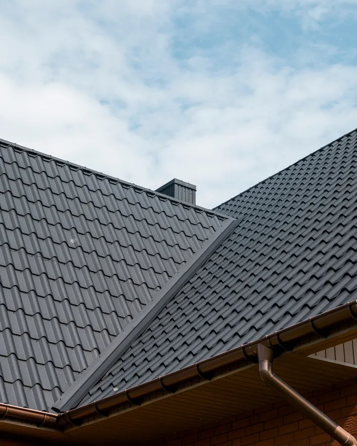 Dark grey tiled roof on a red-brick house, two slopes meeting in a valley above brown guttering, under a bright sky
