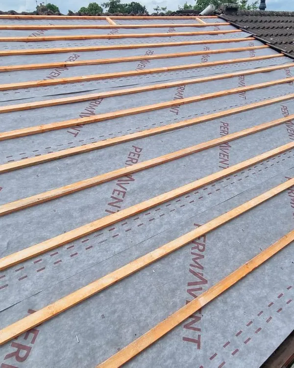 A stripped roof slope covered in grey breathable membrane and rows of new timber battens, next to a neighbour’s old tiled roof