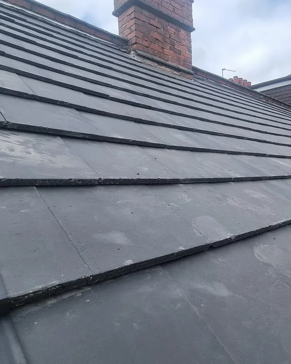 Close view along a new grey tiled roof from the scaffold, showing capped verge tiles at the edge, a black eaves guard above the gutter and a brick chimney