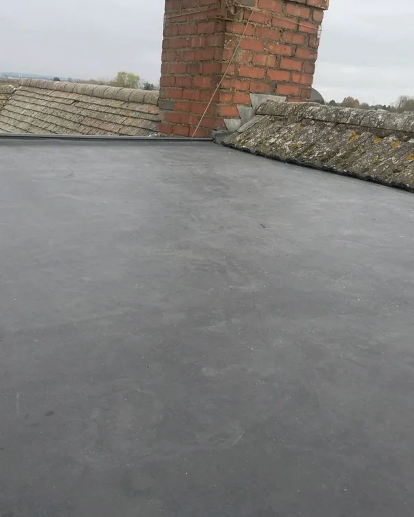 A dark grey rubber flat roof with a neat raised edge, between two lichen-covered tiled slopes and a red-brick chimney