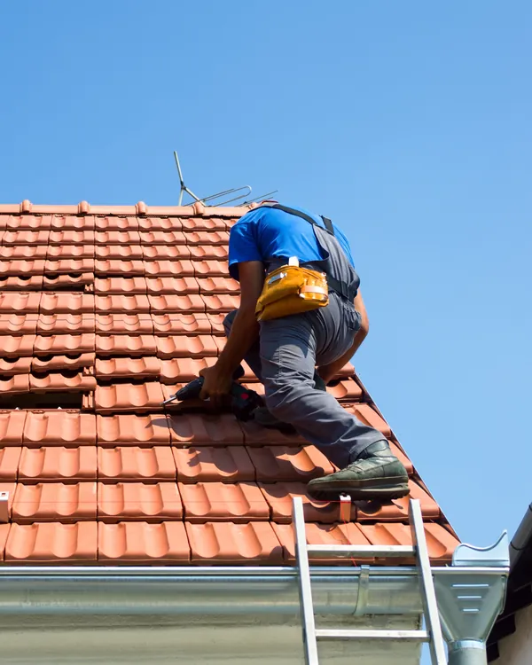 A roofer in a blue T-shirt and tool belt kneels on a red clay tiled roof, refitting lifted tiles beside a ladder