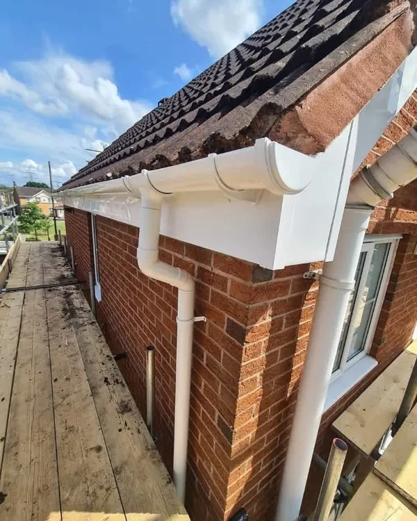 New white uPVC fascia boards, guttering and downpipes on a red-brick house with a brown tiled roof, seen from the scaffold boards